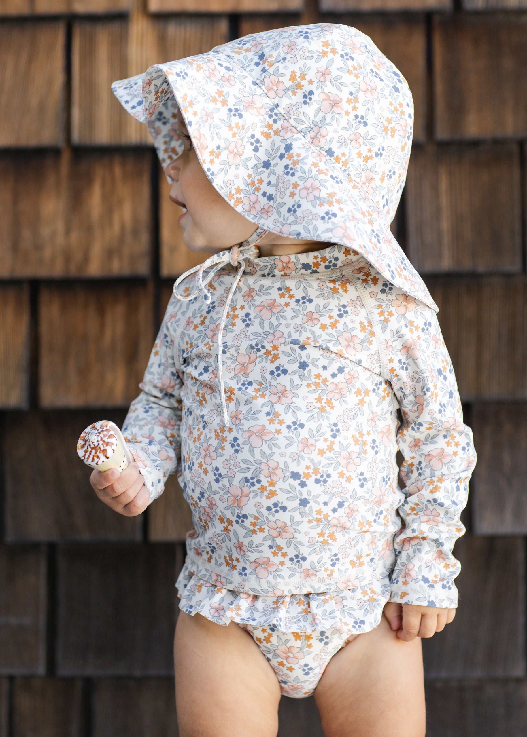 Child wearing a floral sun hat and swimsuit against a wooden wall.