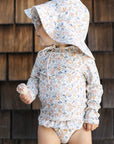 Child wearing a floral sun hat and swimsuit against a wooden wall.
