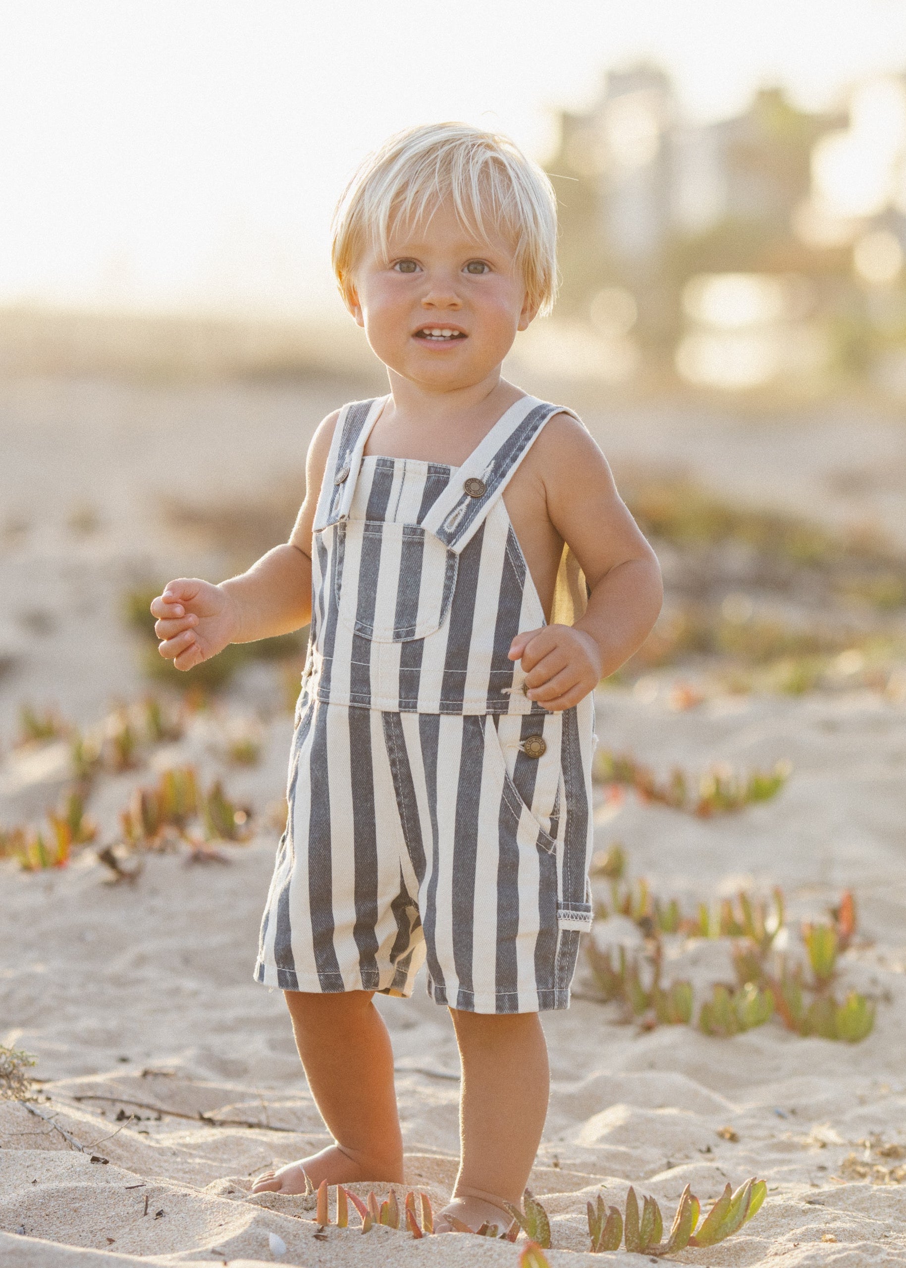 Child wearing striped overalls standing on a sandy beach with a blurred background