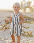 Child wearing striped overalls standing on a sandy beach with a blurred background