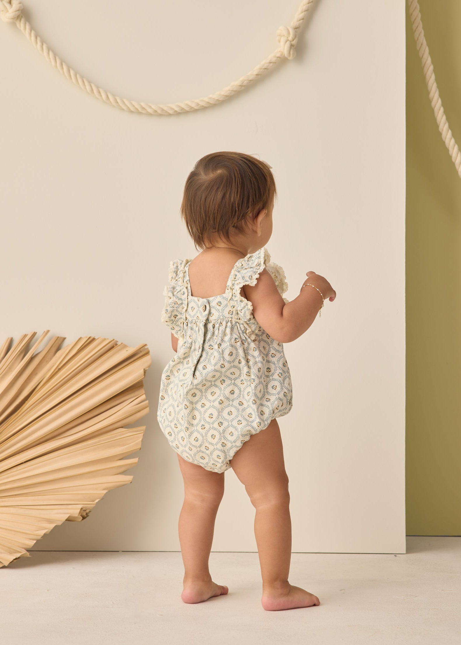 Baby in a floral romper standing against a neutral background with decorative elements.