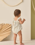 Baby in a floral romper standing against a neutral background with decorative elements.