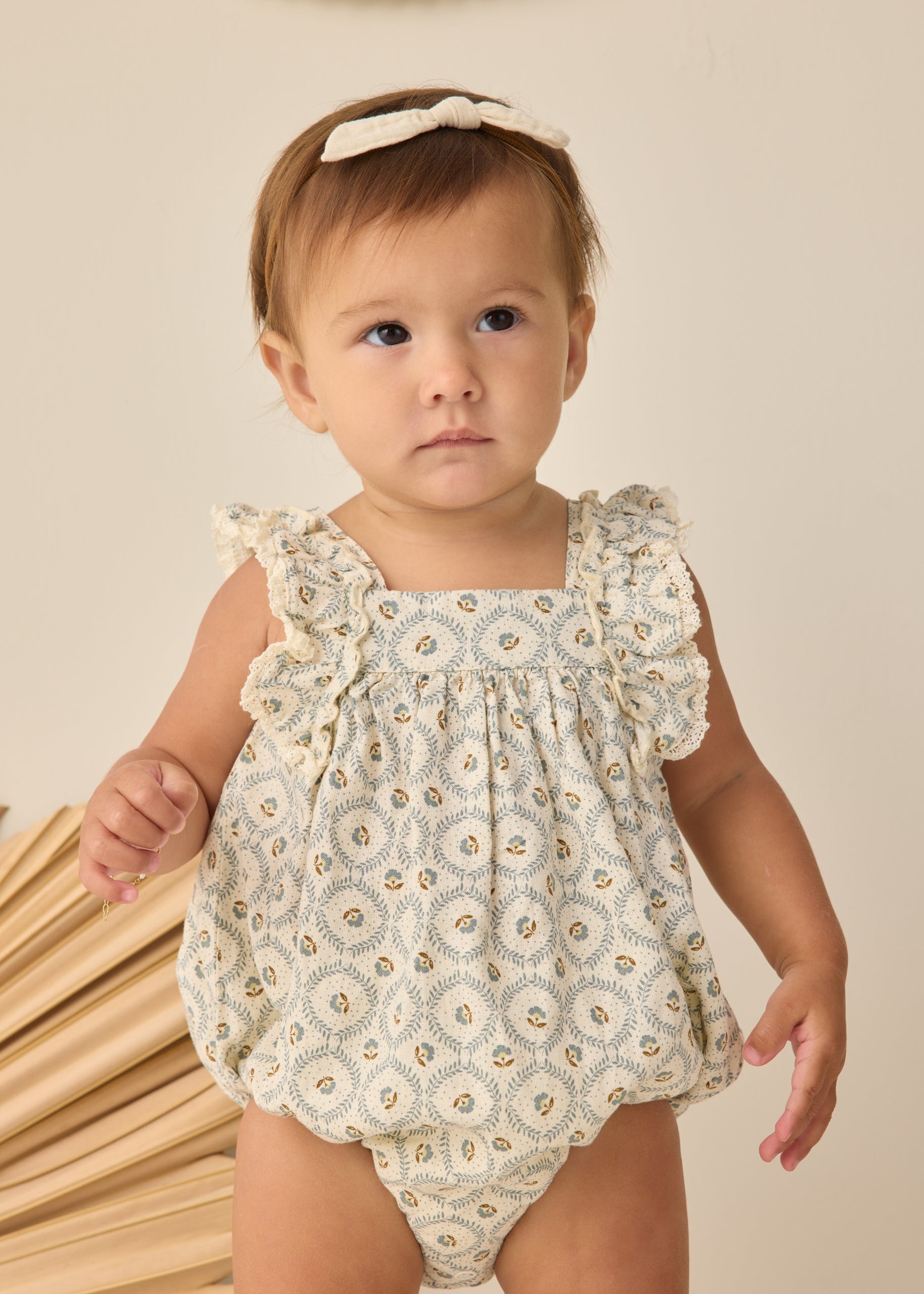 Baby wearing a floral romper standing next to wooden sticks on a beige background