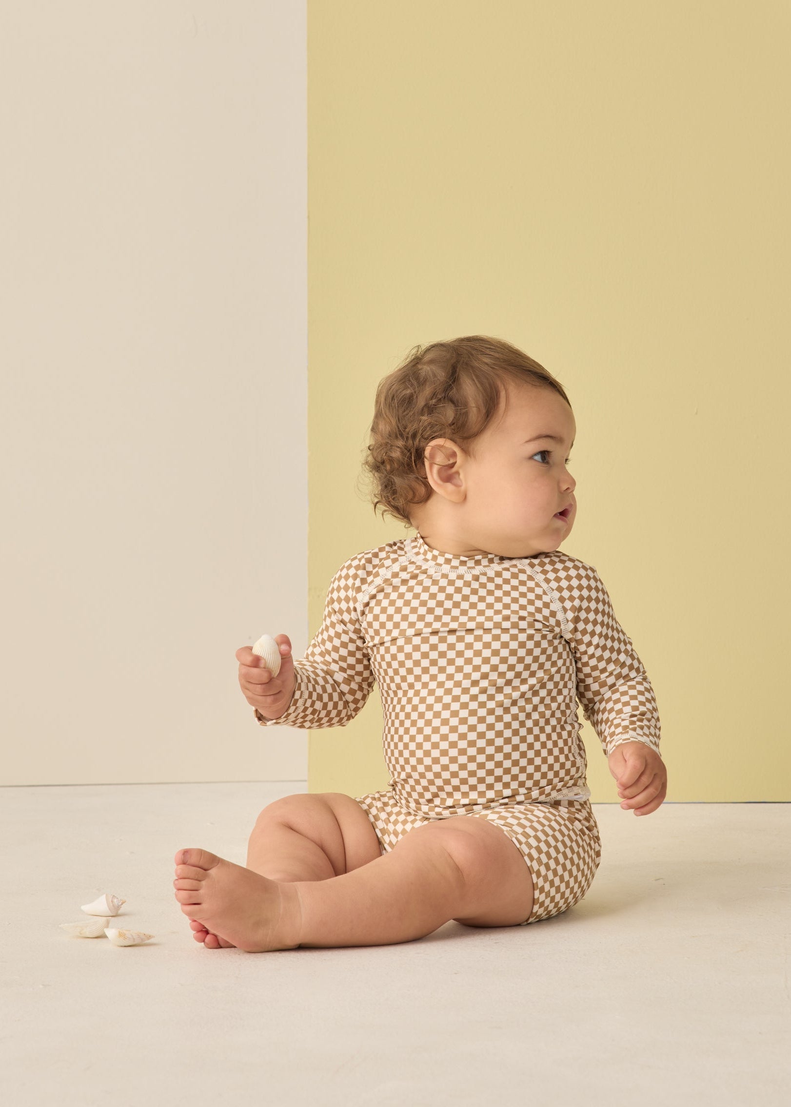 Baby sitting on a light-colored floor wearing a checkered outfit against a yellow wall.