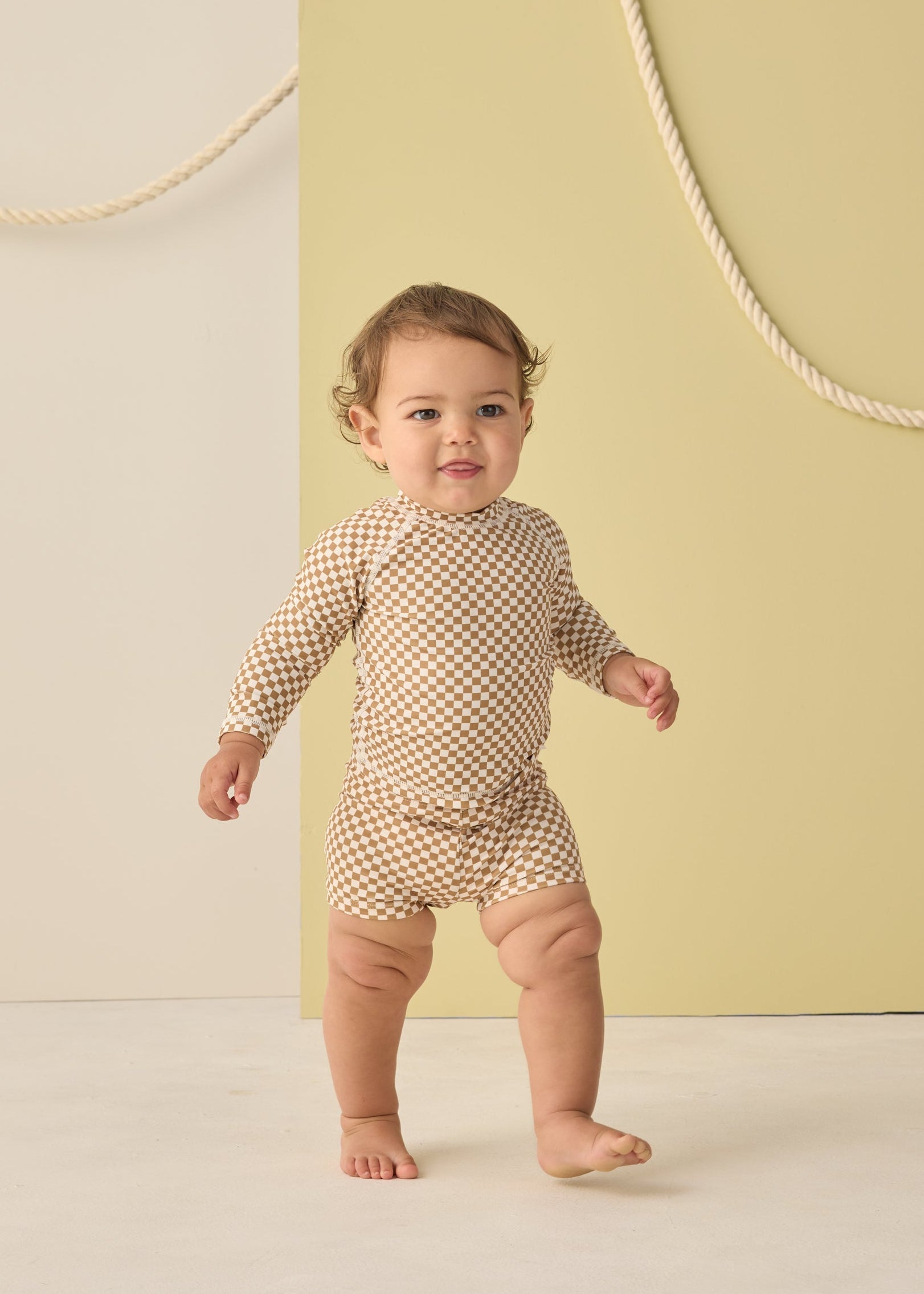 Baby standing in a checkered outfit against a light yellow background