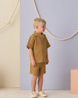 Child wearing a mustard checkered outfit standing against a light purple wall with decorative ropes.