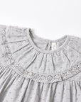 Close-up of a light gray baby dress featuring a textured fabric, a large pleated ruffled collar with delicate lace trim, and a small bow pattern.