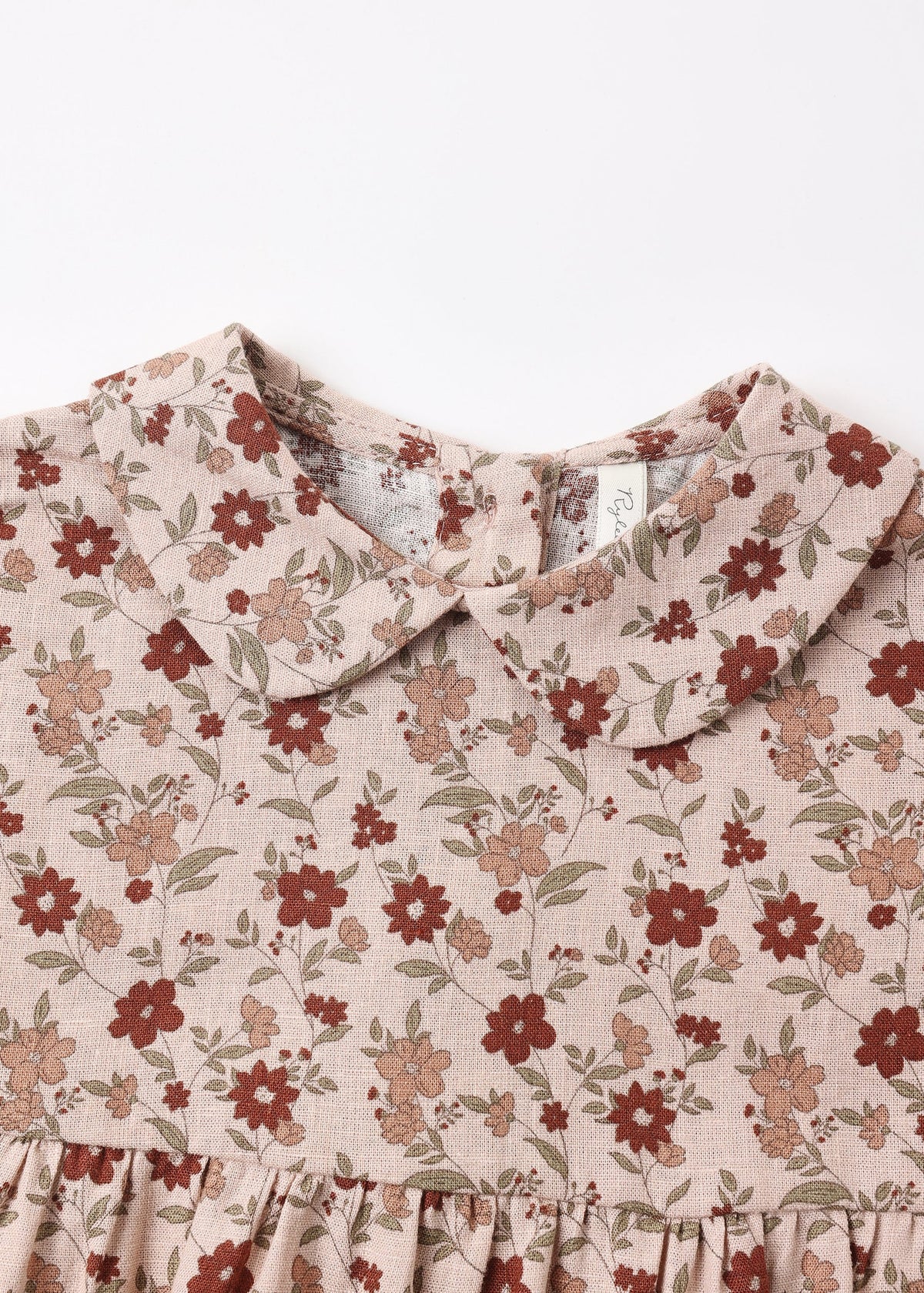 Close-up of a children's dress with a ruffled Peter Pan collar and a dusty floral print on a light beige linen-like fabric, featuring red and brown flowers with green leaves.