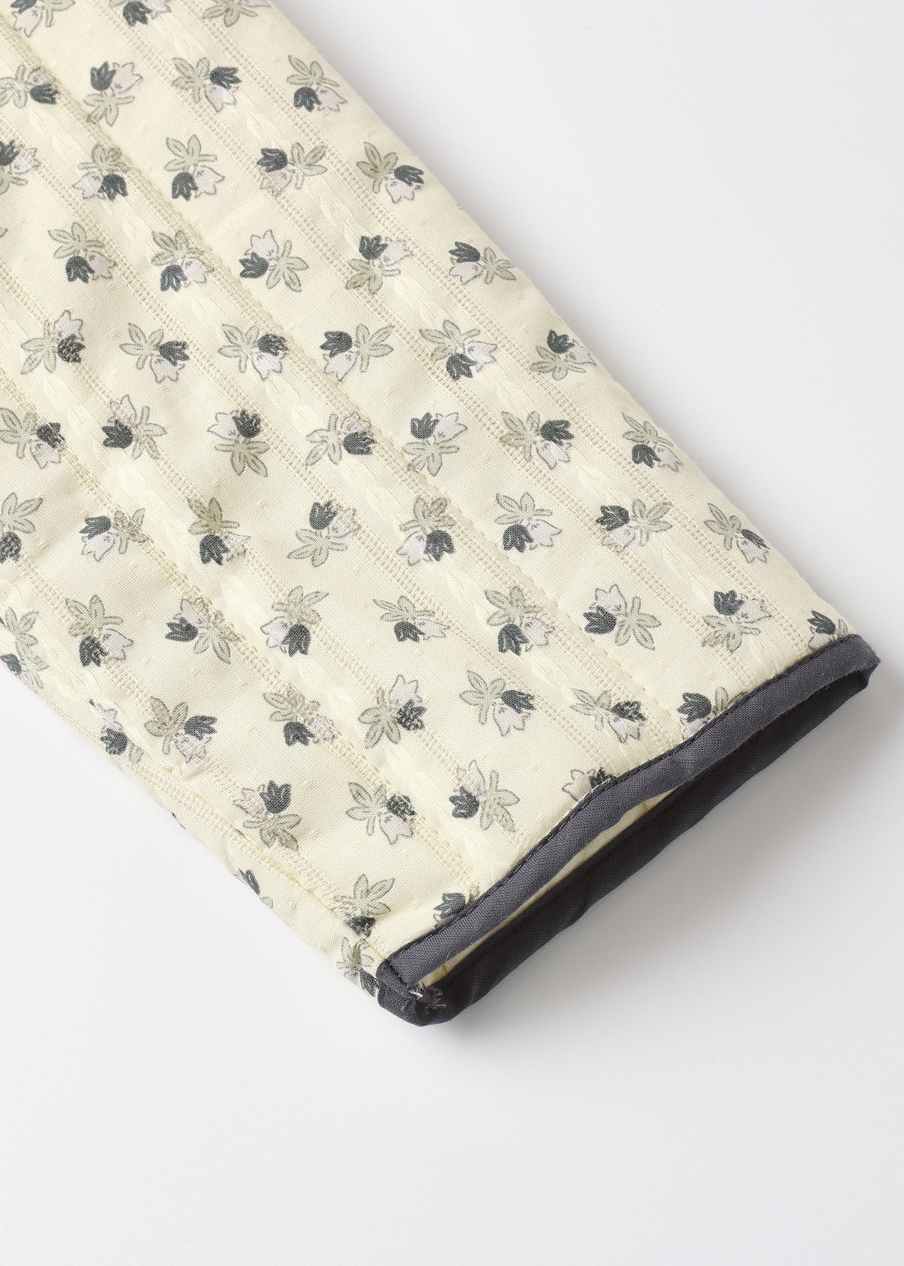 A close-up of a quilted blanket or comforter with a cream-colored background and a repeating pattern of small gray and black floral motifs, finished with a dark gray piped edge.
