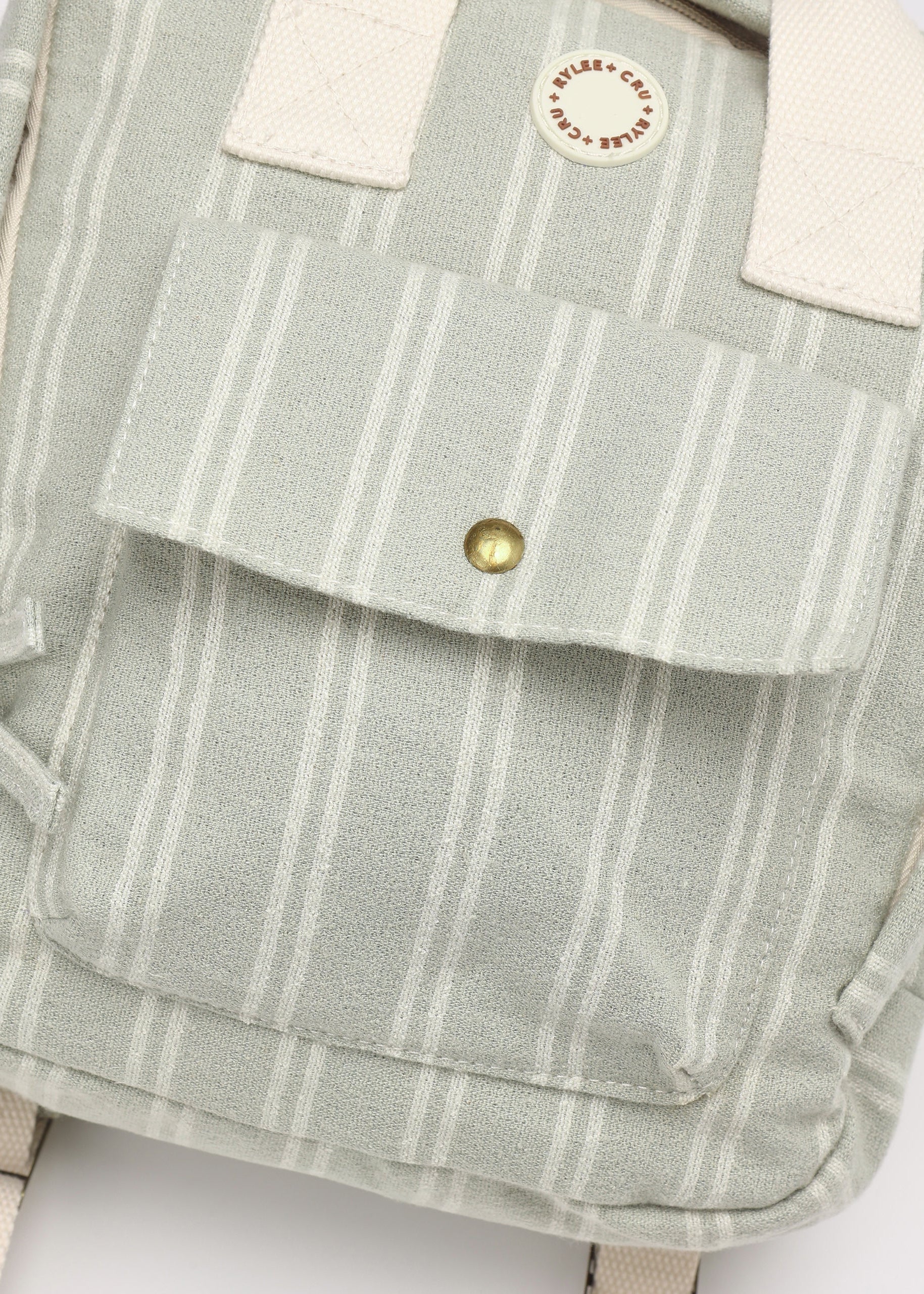 Close-up of a canvas backpack in a pale sage green and white stripe pattern, featuring a front pocket with a brass snap button, off-white webbing straps, and a circular brand patch reading 'MILKY CAKE TOYS'.