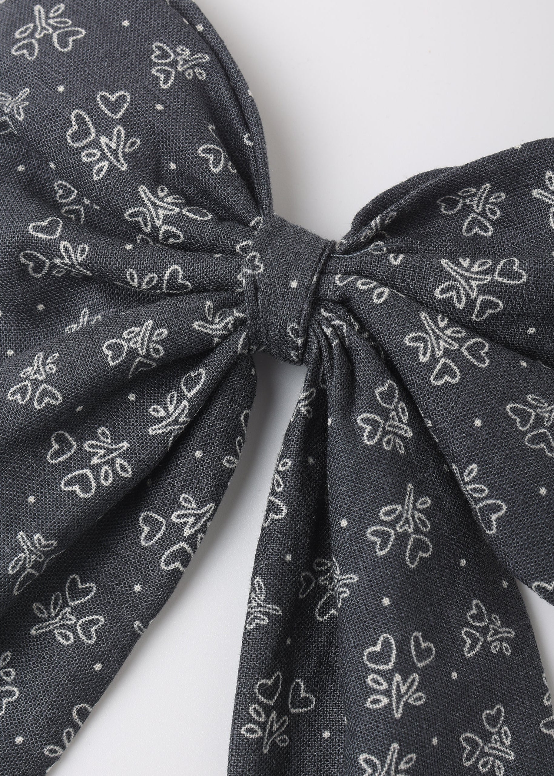 A close-up of a dark gray fabric bow with a white line-art pattern of small hearts and floral motifs, set against a plain light gray background.