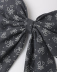 A close-up of a dark gray fabric bow with a white line-art pattern of small hearts and floral motifs, set against a plain light gray background.