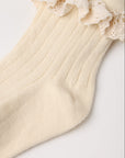 A close-up of a cream-colored ribbed knit sock with a matching lace ruffle trim at the top, laid flat on a light background.