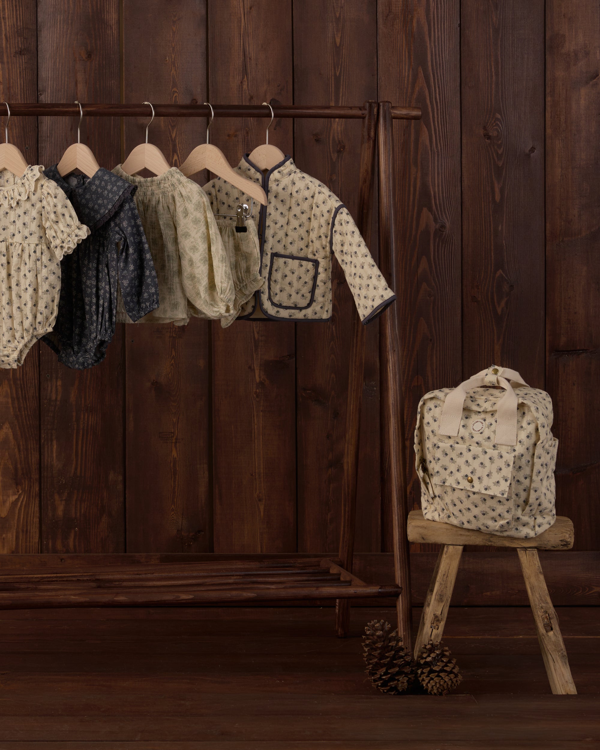 A set of children's patterned knit clothing displayed on a wooden hanger rack against a dark wood-paneled wall, with a matching cream patterned tote bag resting on a small wooden stool.