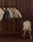 A set of children's patterned knit clothing displayed on a wooden hanger rack against a dark wood-paneled wall, with a matching cream patterned tote bag resting on a small wooden stool.