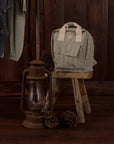 A light gray and white vertically striped canvas backpack with cream-colored top handles, displayed on a rustic wooden stool next to an antique-style lantern and pinecones against a dark wood-paneled background.