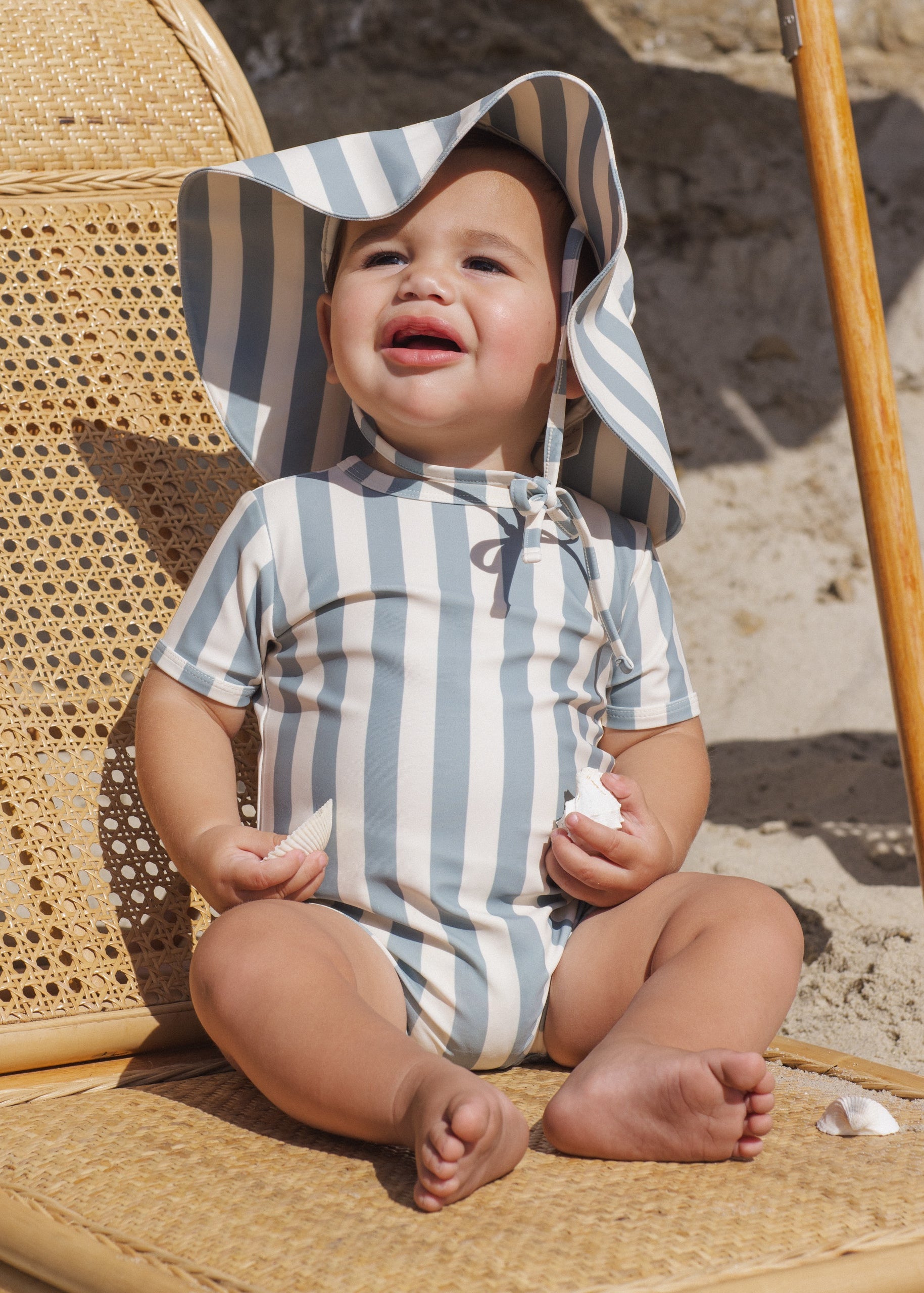 Baby in a striped swimsuit and sun hat sitting on a sandy beach.