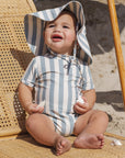 Baby in a striped swimsuit and sun hat sitting on a sandy beach.
