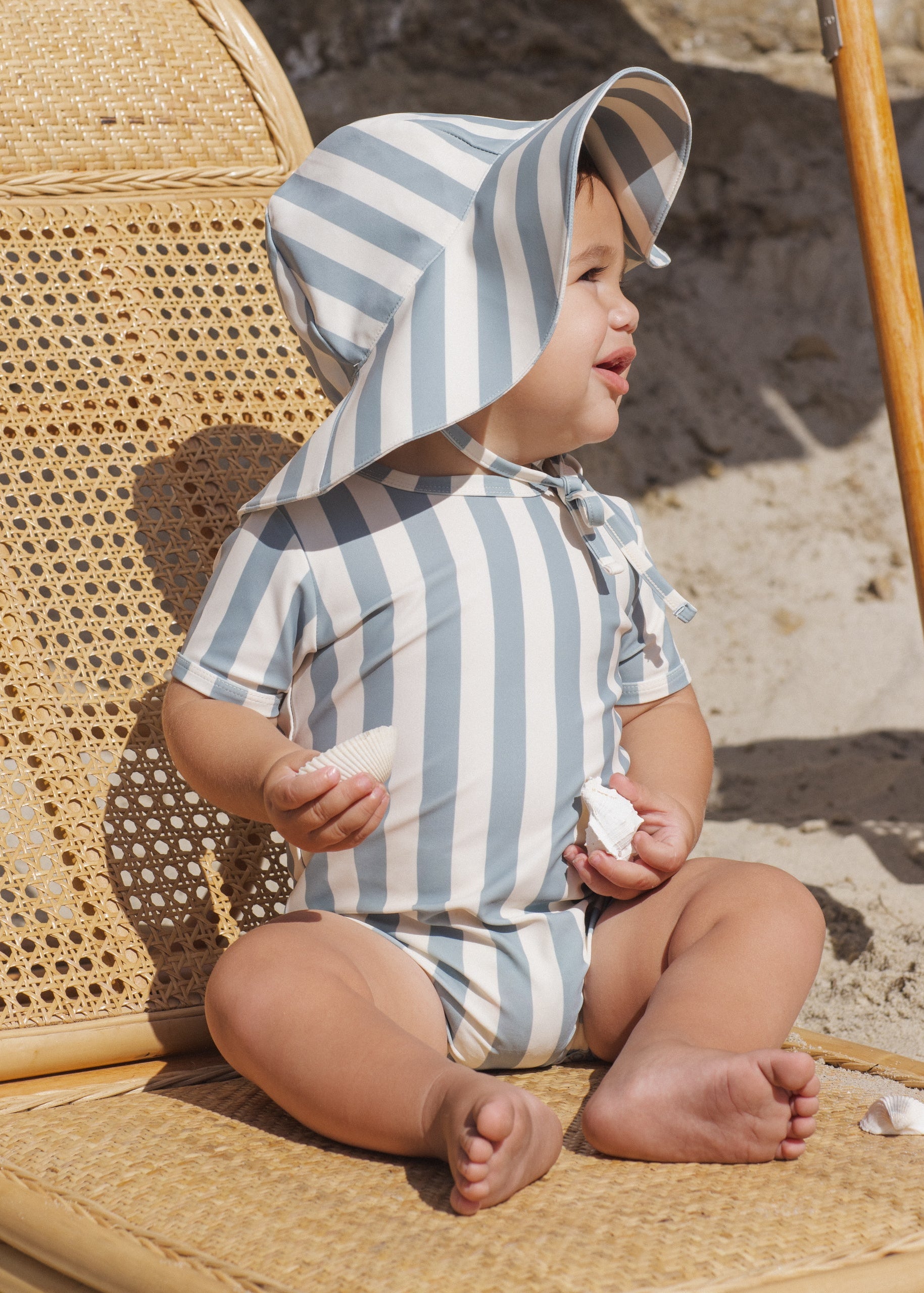 Baby wearing a striped sun hat and swimsuit sitting on a woven chair.