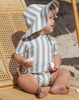 Baby wearing a striped sun hat and swimsuit sitting on a woven chair.