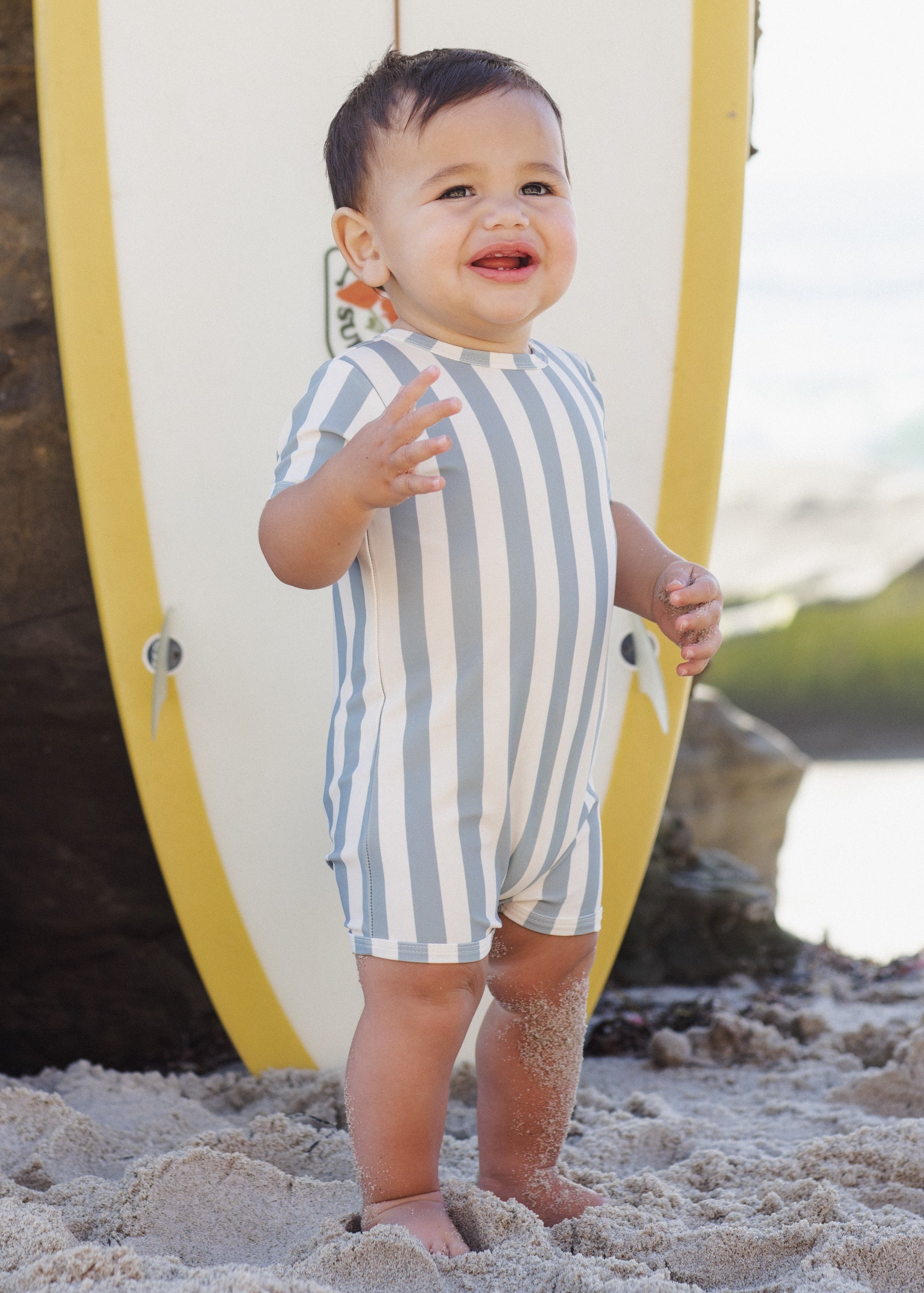 Child in a striped romper standing on a beach with a surfboard in the background