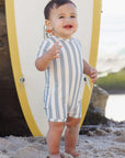 Child in a striped romper standing on a beach with a surfboard in the background