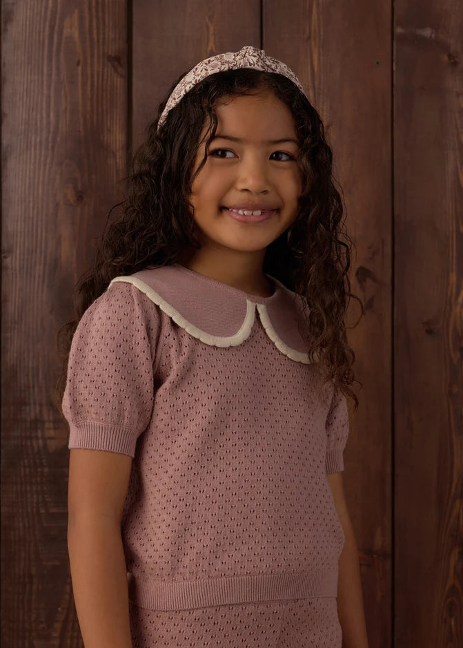 A young girl with long curly brown hair wearing a dusty pink knit top with a cream Peter Pan collar and a matching patterned headband, standing against a dark wood plank background.