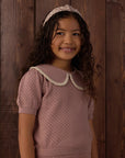 A young girl with long curly brown hair wearing a dusty pink knit top with a cream Peter Pan collar and a matching patterned headband, standing against a dark wood plank background.