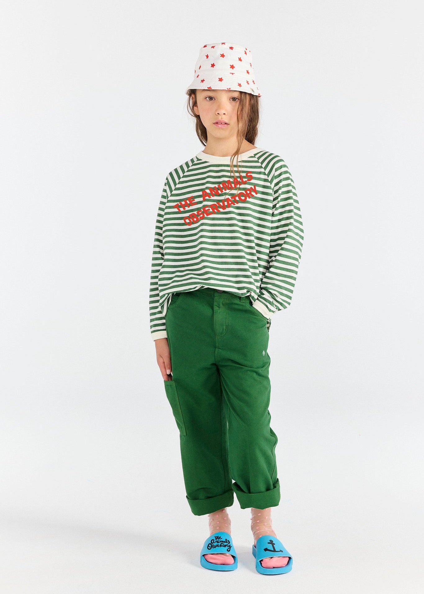 Person wearing a green and white striped shirt with red text, green pants, and blue sandals on a white background.