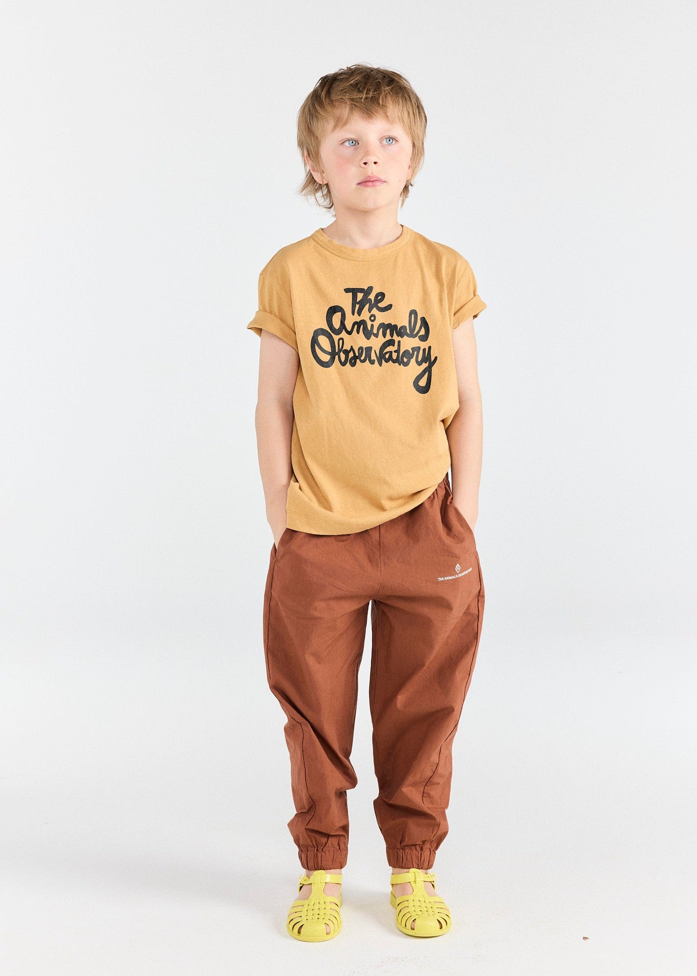 Child wearing a mustard yellow t-shirt with text and brown pants on a white background
