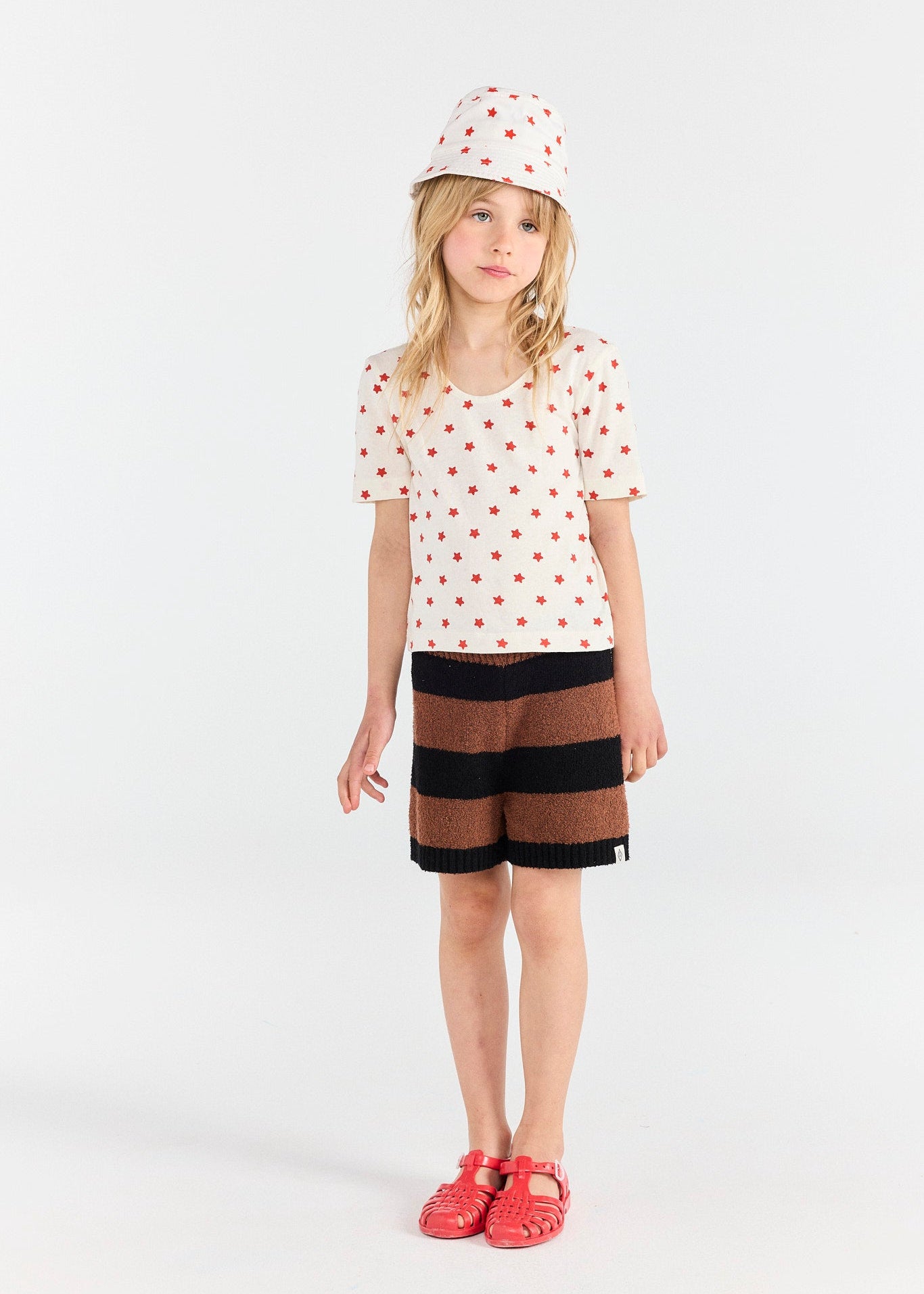 Young girl wearing a polka dot shirt, striped shorts, and red shoes on a white background