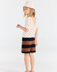 Young girl wearing a white polka dot dress with a black and brown striped skirt on a white background