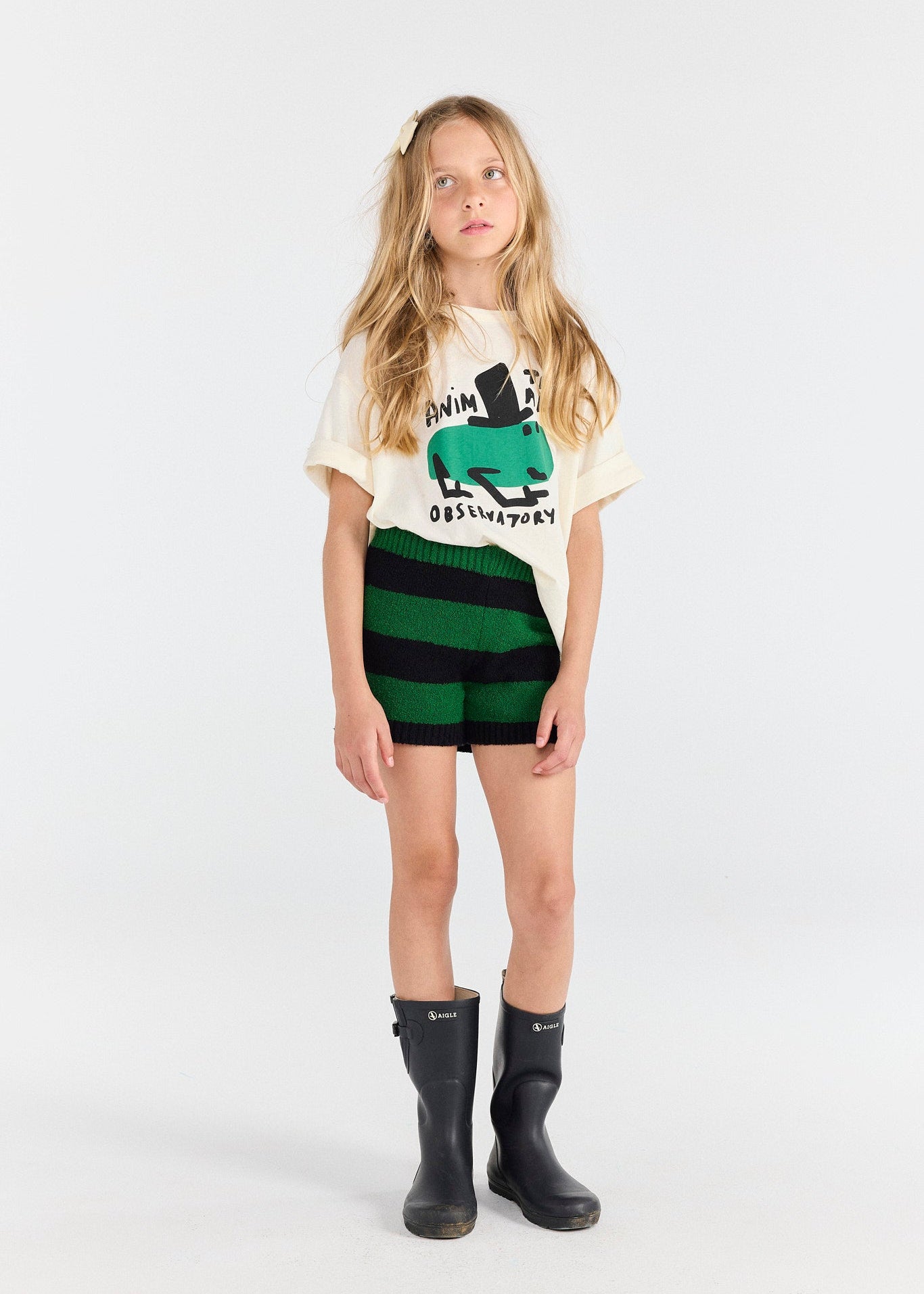 Young girl wearing a graphic t-shirt, striped shorts, and rain boots on a white background