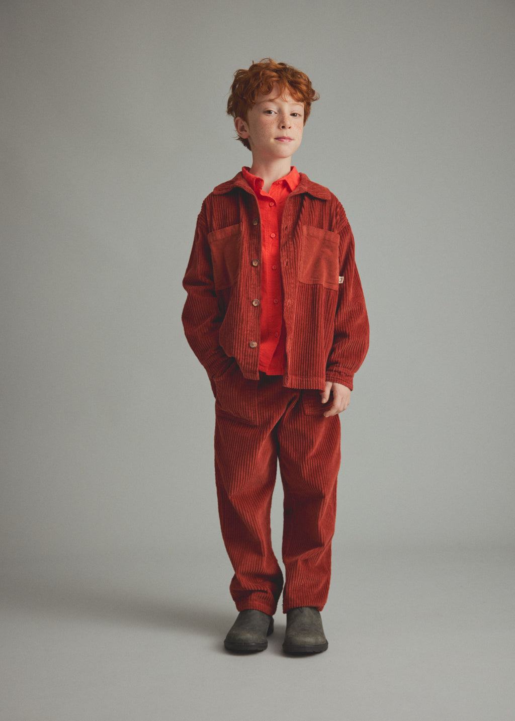 A young boy with red hair stands against a neutral gray studio background wearing a rust-orange corduroy jacket and matching corduroy pants over a bright red collared shirt, with dark gray boots.