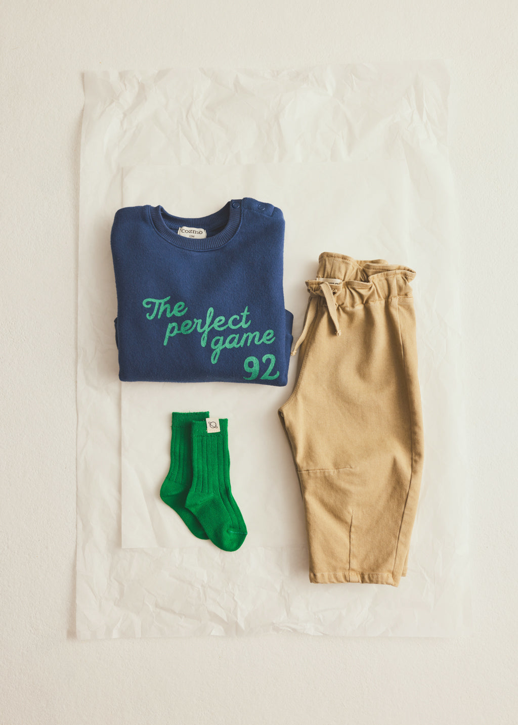 A flat-lay of a folded navy blue sweatshirt with green text reading 'The perfect game 92', paired with tan pants and bright green socks, arranged on a white sheet of paper.