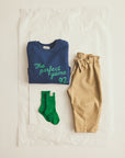 A flat-lay of a folded navy blue sweatshirt with green text reading 'The perfect game 92', paired with tan pants and bright green socks, arranged on a white sheet of paper.