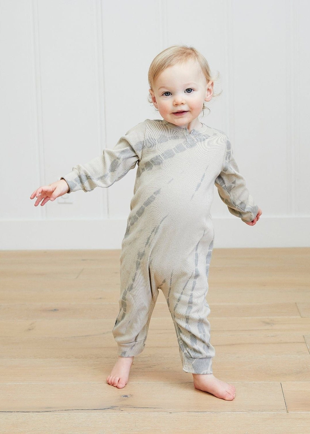 Child wearing a gray and white striped onesie standing on a wooden floor.