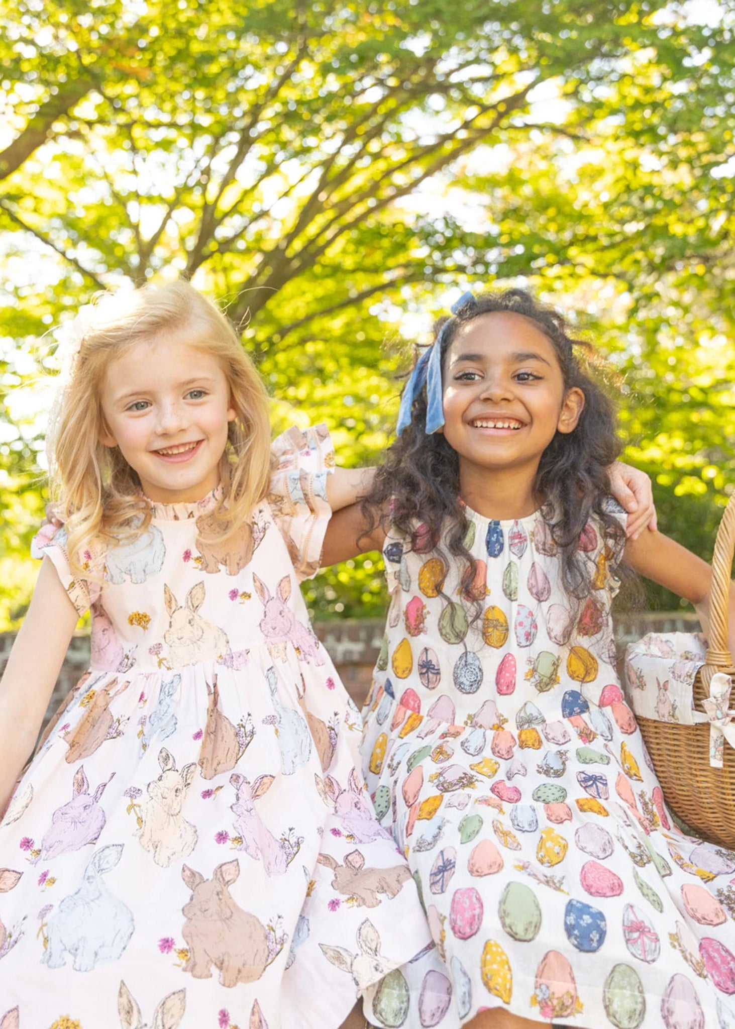 Two young girls in patterned dresses standing outdoors with a picnic basket.