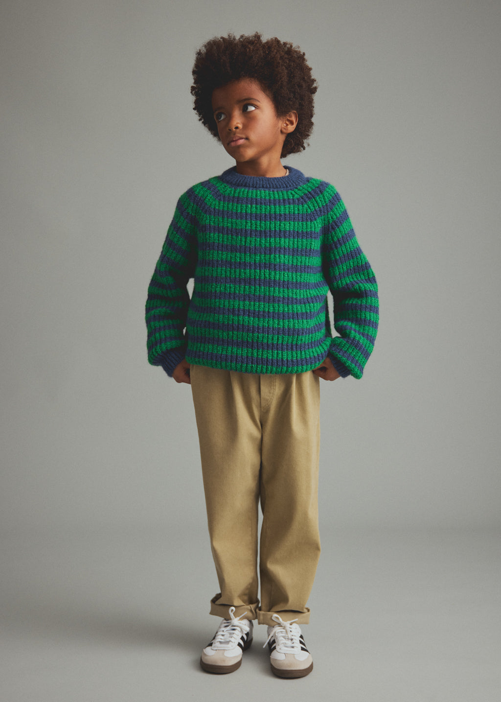 A child model wearing a green and dark blue striped knit sweater, beige trousers, and white sneakers, standing against a neutral gray studio background.
