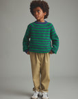 A child model wearing a green and dark blue striped knit sweater, beige trousers, and white sneakers, standing against a neutral gray studio background.