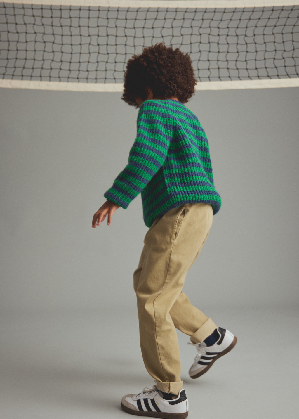 A child wearing a green and dark striped knit sweater, beige pants, and white sneakers with black stripes, standing in a studio with a gray background and a tennis net above.