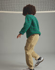 A child wearing a green and dark striped knit sweater, beige pants, and white sneakers with black stripes, standing in a studio with a gray background and a tennis net above.