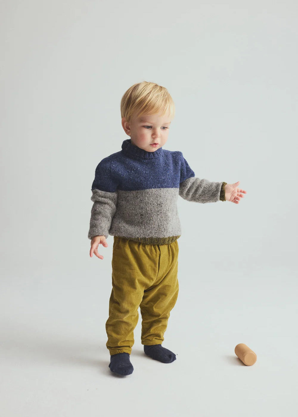 A toddler standing in a studio wearing a color-block knit sweater with navy blue, gray, and beige sections, olive yellow pants, and dark socks. A small wooden toy lies on the floor nearby against a light gray background.