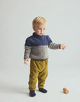 A toddler standing in a studio wearing a color-block knit sweater with navy blue, gray, and beige sections, olive yellow pants, and dark socks. A small wooden toy lies on the floor nearby against a light gray background.