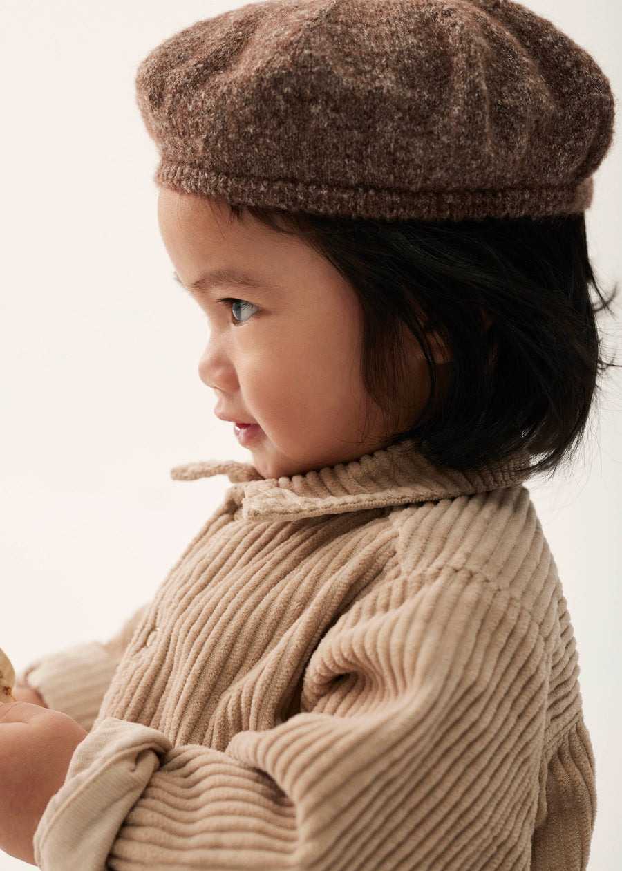 Child wearing a brown knitted hat and beige sweater against a neutral background
