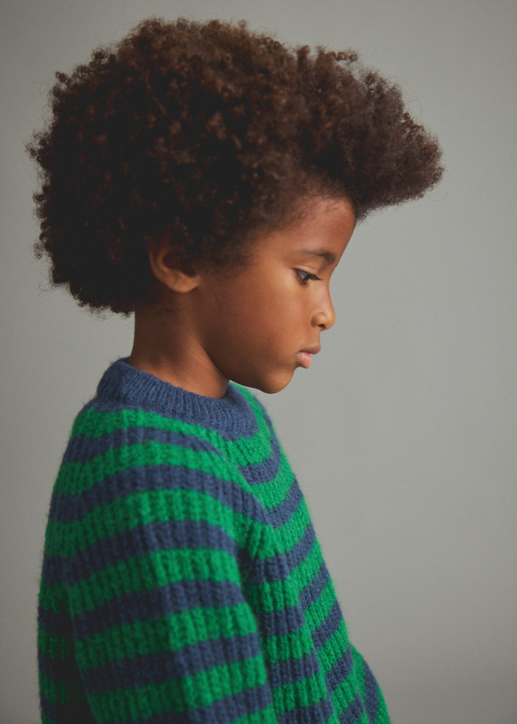 A child with voluminous dark brown curly hair shown in profile, wearing a chunky knit sweater with horizontal stripes in green and navy blue against a plain gray studio background.
