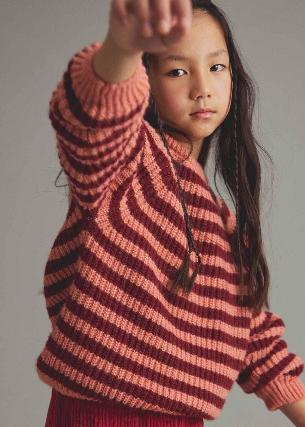 A young girl with long dark hair wearing a chunky knit striped sweater in shades of dusty pink and deep burgundy, posing with one arm raised against a plain gray studio background.