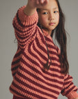 A young girl with long dark hair wearing a chunky knit striped sweater in shades of dusty pink and deep burgundy, posing with one arm raised against a plain gray studio background.