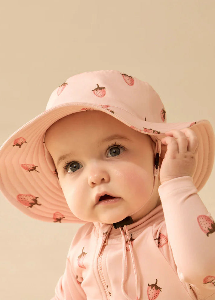 Baby wearing a pink sun hat and matching outfit with strawberry pattern on a beige background