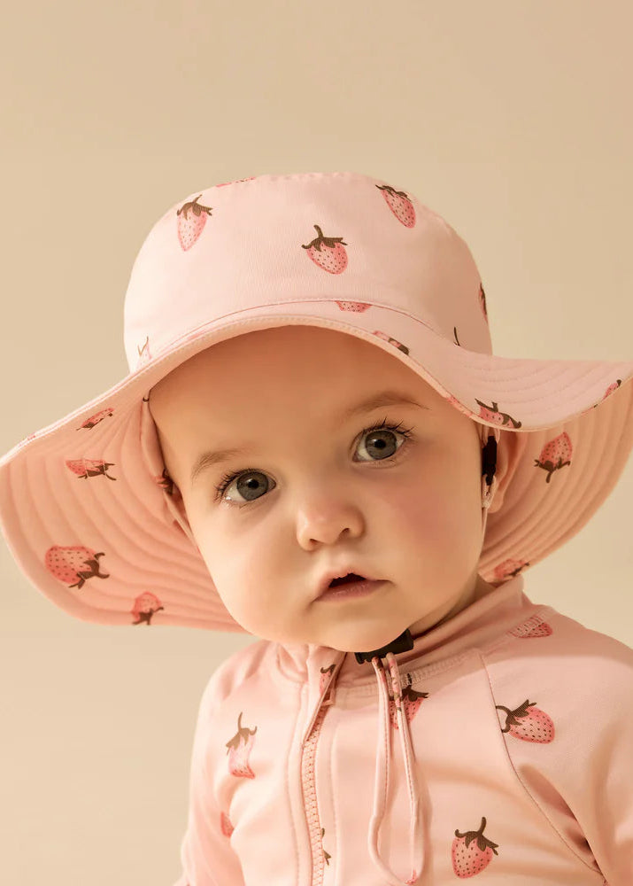 Child wearing a pink sun hat and jacket with strawberry pattern on a beige background